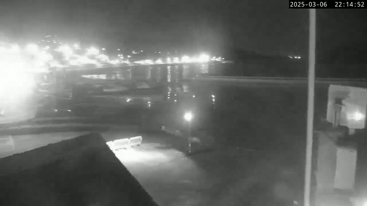 A nighttime, low-light view shows a dimly lit area with a body of water reflecting lights in the distance and a vehicle parked nearby.