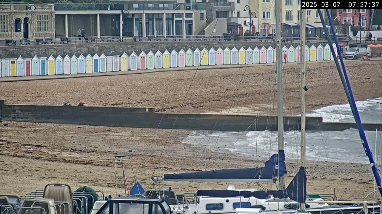 A row of colorful beach huts lines a pebble beach with gentle waves lapping the shore, and several boats are moored nearby.