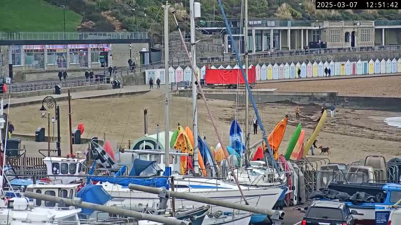 A harbor filled with boats and kayaks sits on a sandy beach next to a row of colorful beach huts, under an overcast sky.