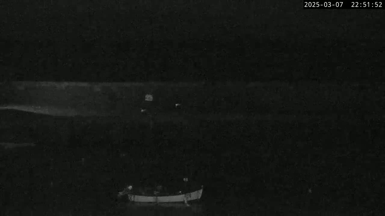 A low-light nighttime image shows a dark body of water under a cloudy sky, with a boat near the bottom of the frame and faint lights visible in the distance.