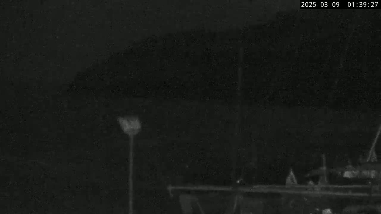 A nighttime, low-light image shows a dark, indistinct landscape with a single light pole and a structure that appears to be a boat or dock in the bottom right corner.