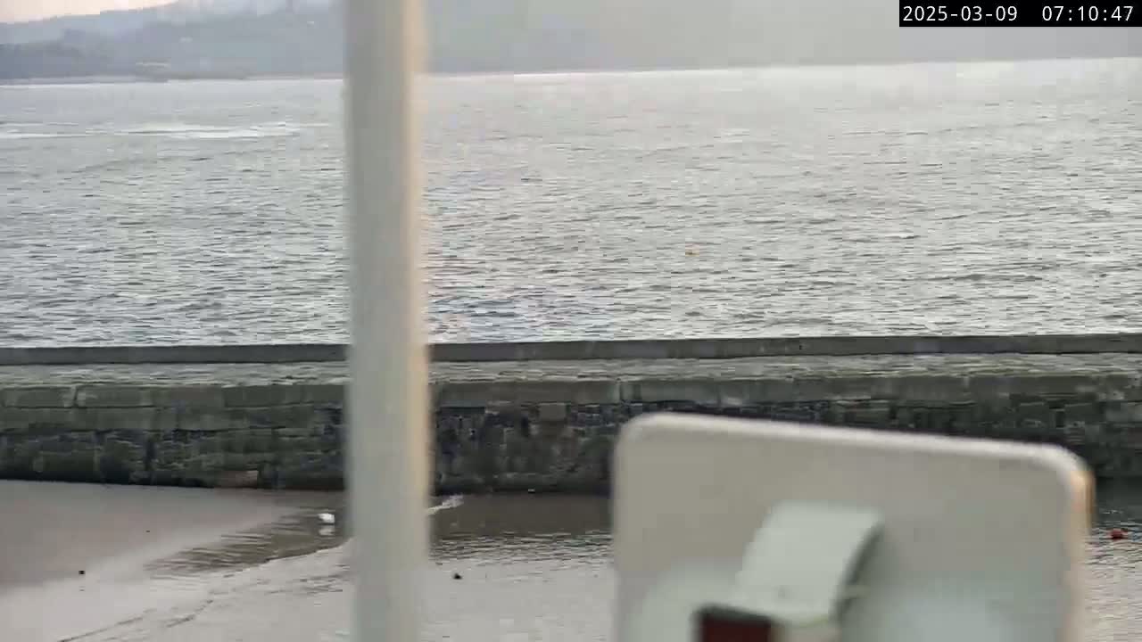 A somewhat choppy body of water is seen beyond a low stone wall, under a cloudy sky.