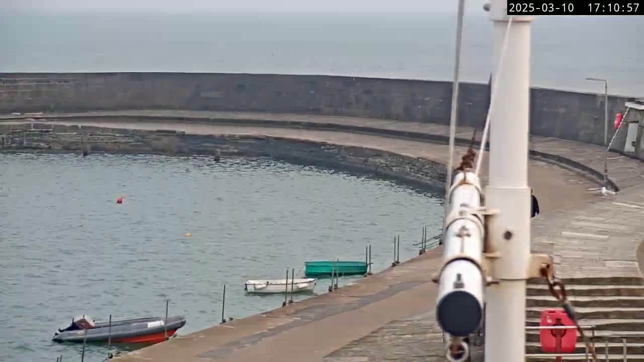 A curved stone harbor wall surrounds a calm body of water containing several small boats on an overcast day.