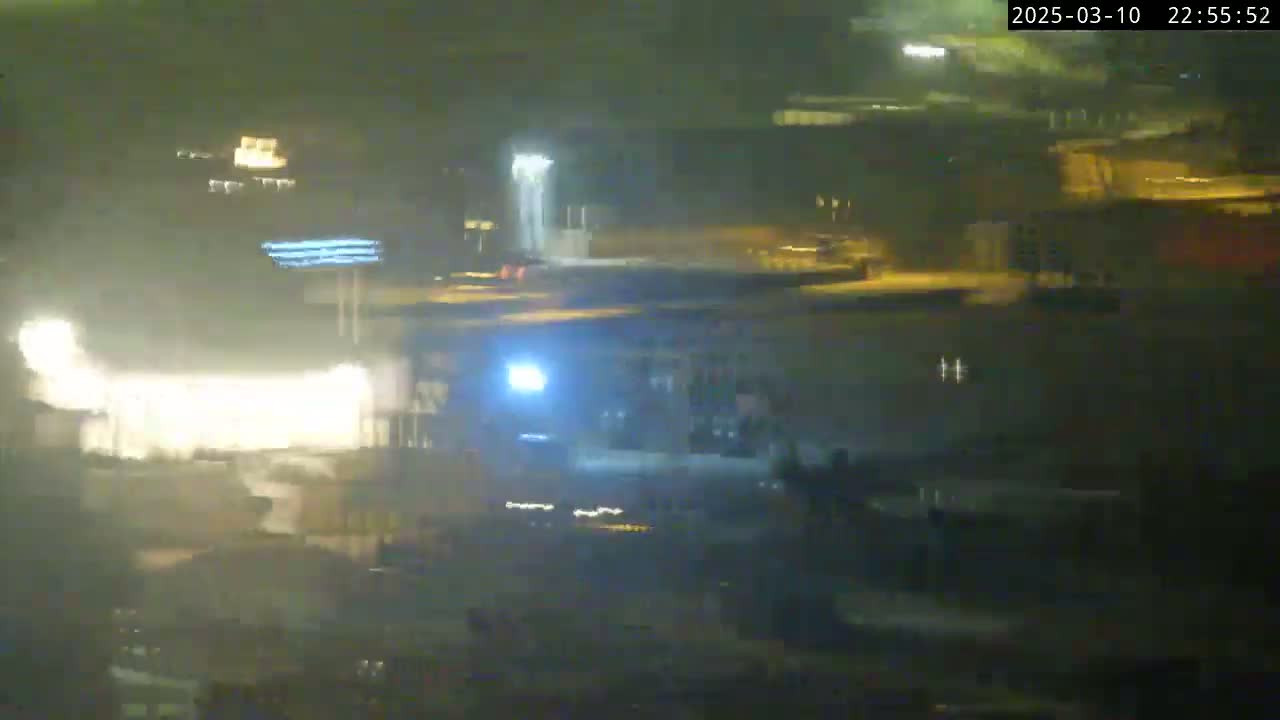 A blurry nighttime view of a dimly lit area with several buildings and vehicles, possibly under rainy conditions.