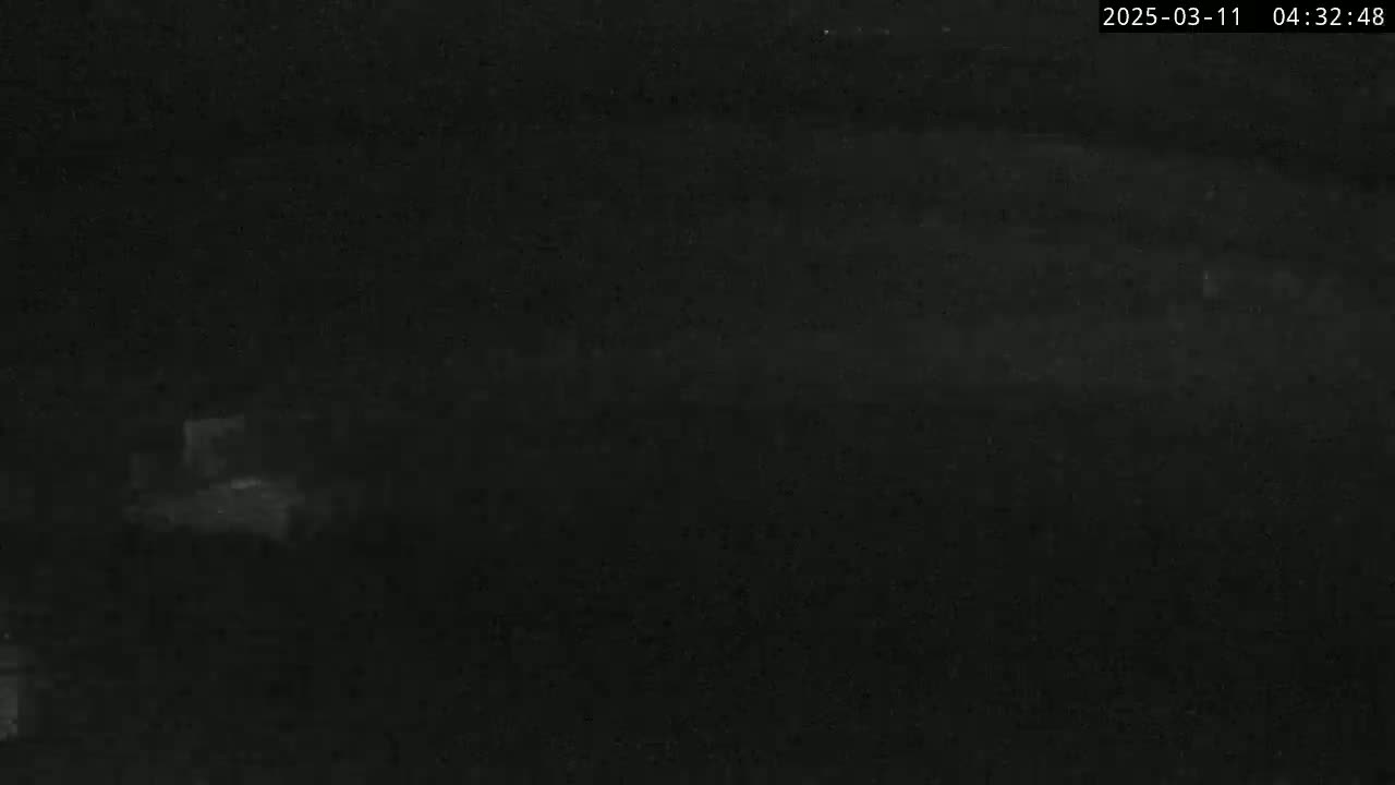A dark, nighttime image shows a dimly lit area with faint shapes suggesting vehicles or structures in the distance.