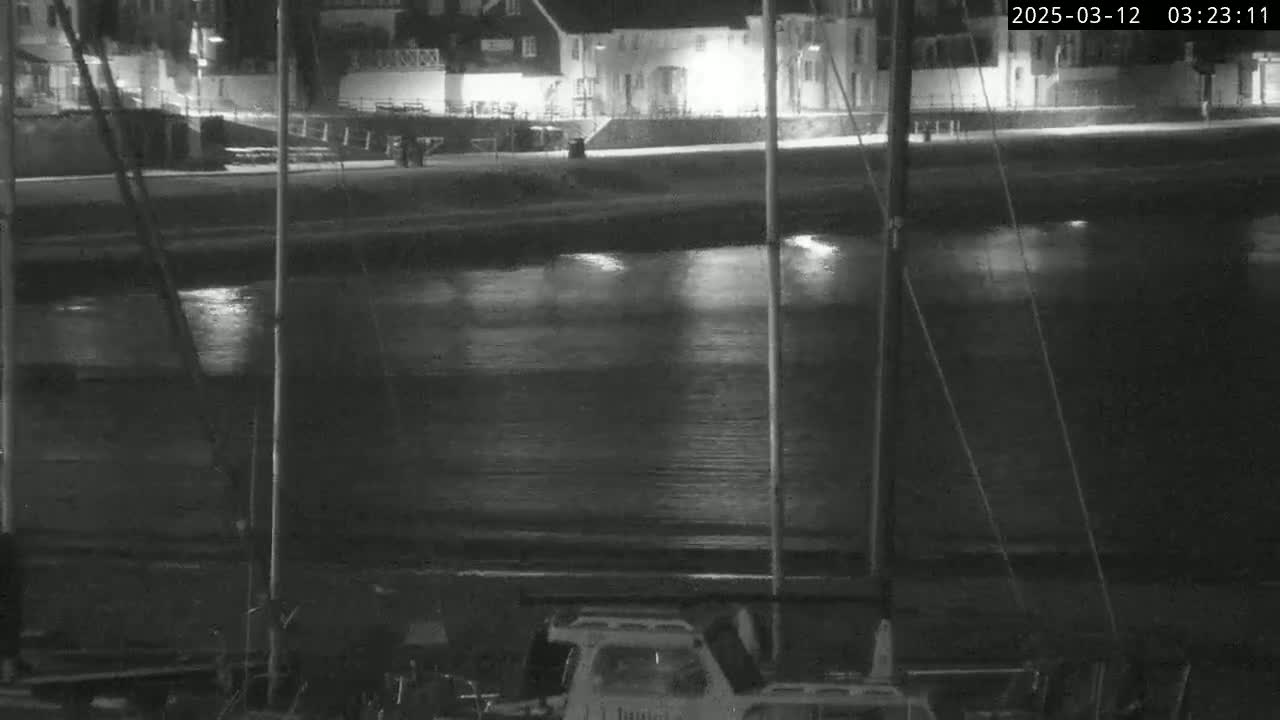 A nighttime view of a calm body of water reflecting lights from buildings on the shore, with several masts of boats visible in the foreground.