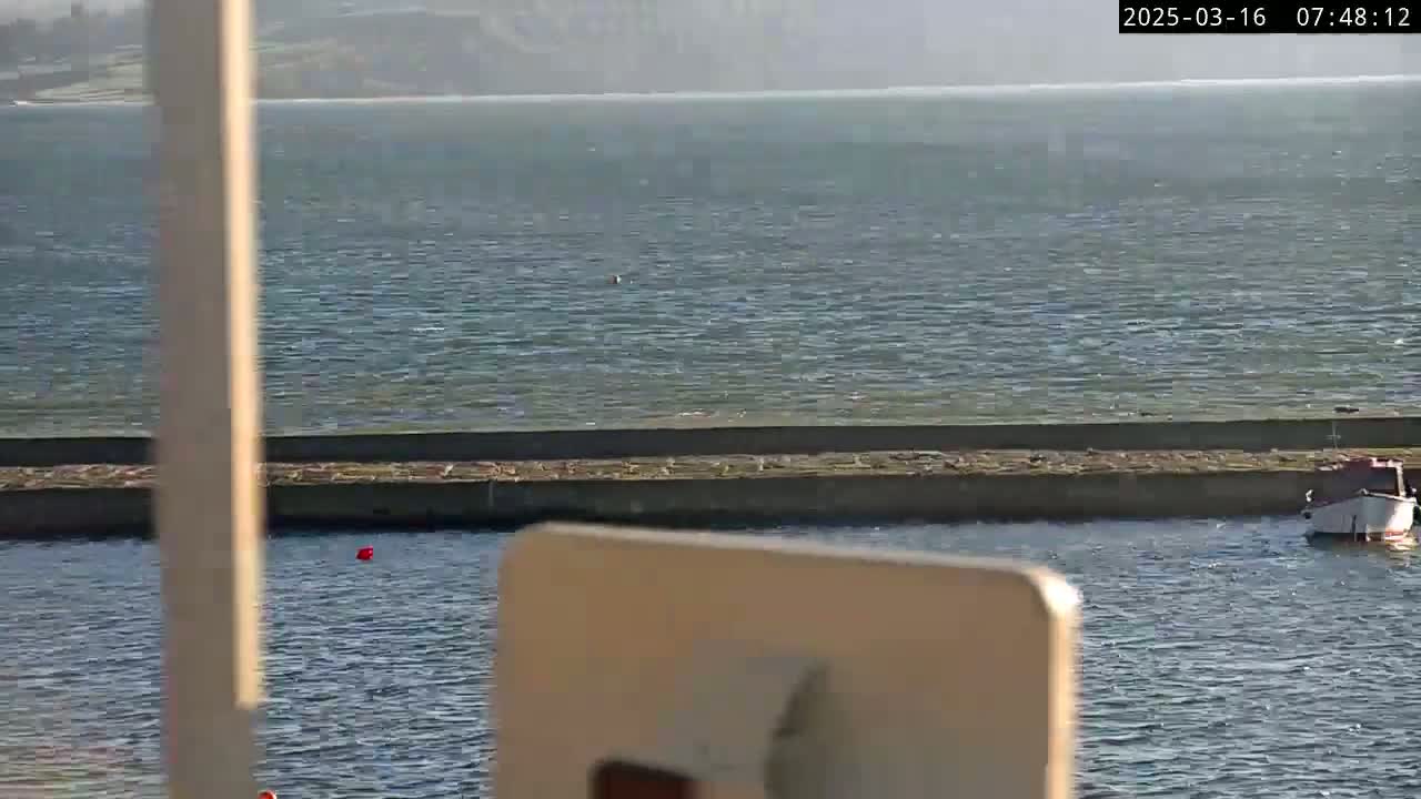 A calm body of water with a small boat and a low stone wall is seen from a partially obstructed view.