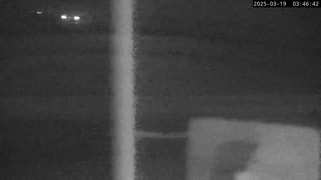 A nighttime, low-light image shows a blurry, indistinct scene possibly of a road with faint headlights in the distance and a vertical pole or post in the foreground.
