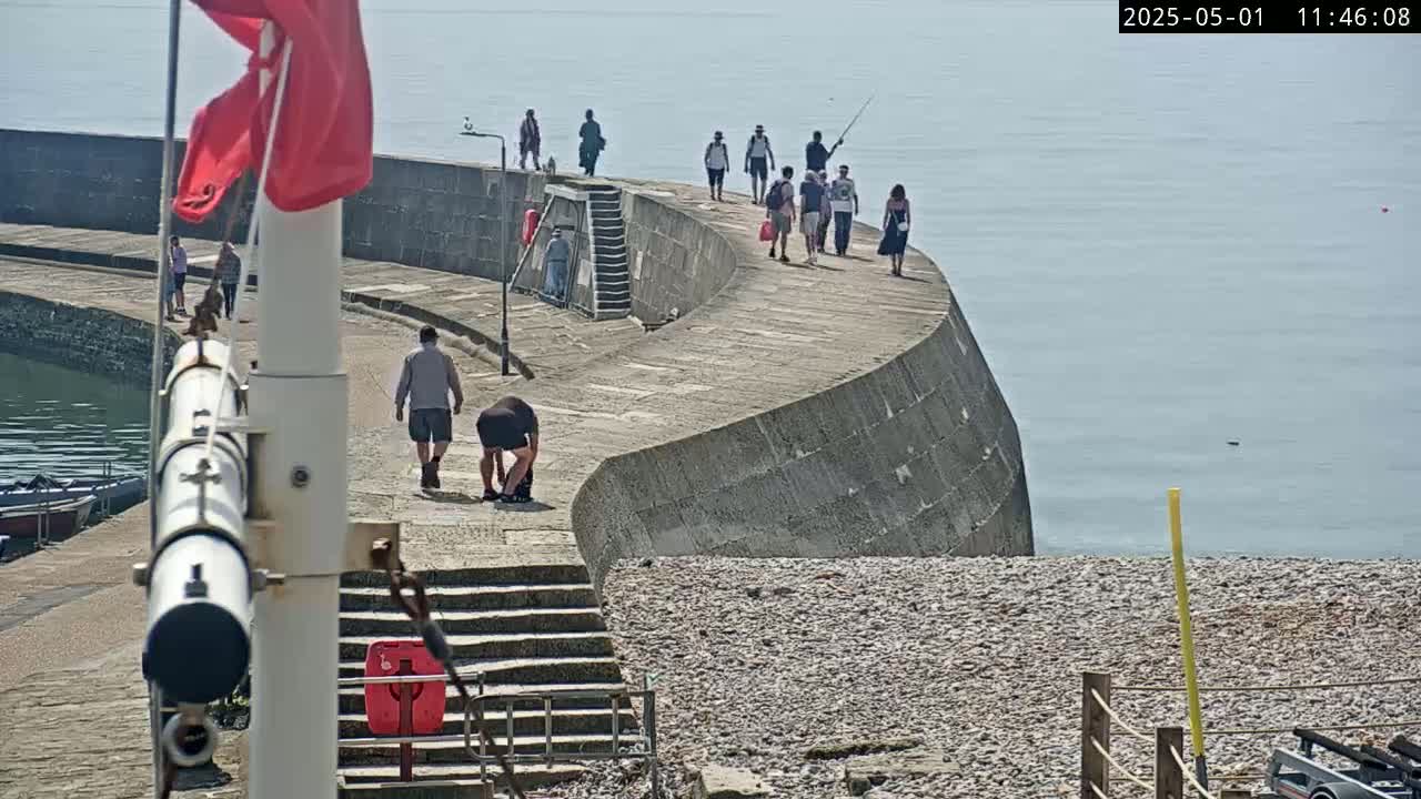 A curved stone wall, overlooking a calm sea on a sunny day, is lined with several people walking and fishing.