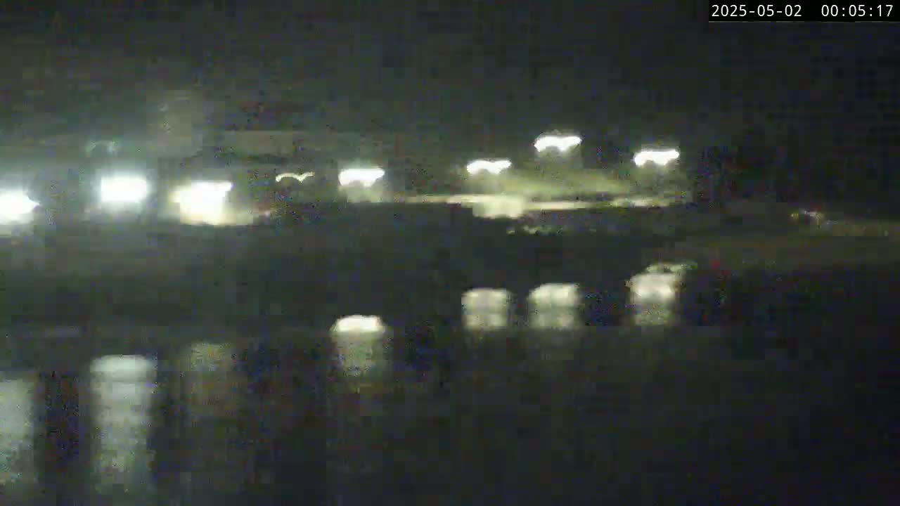 A nighttime, low-light view shows a roadway under a bridge, with vehicle headlights visible in the distance and reflections in the water below.