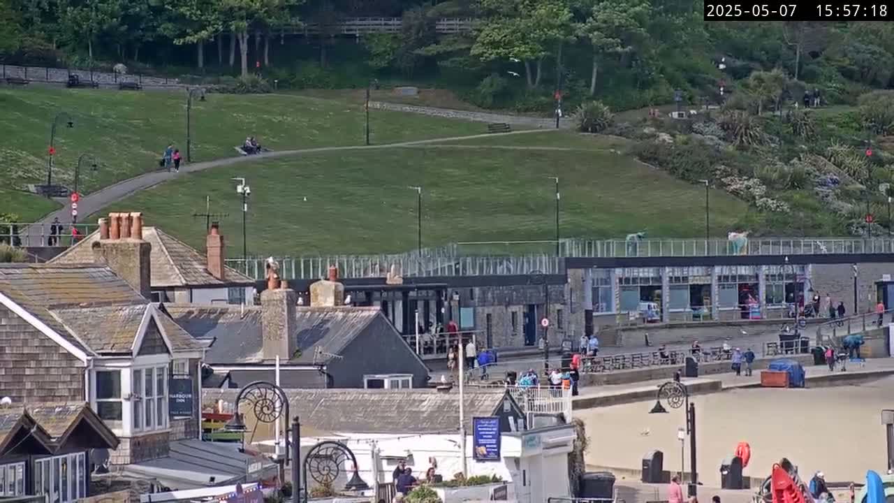 A sunny seaside town scene shows a grassy hill overlooking a beach with buildings, people walking, and various outdoor seating areas.