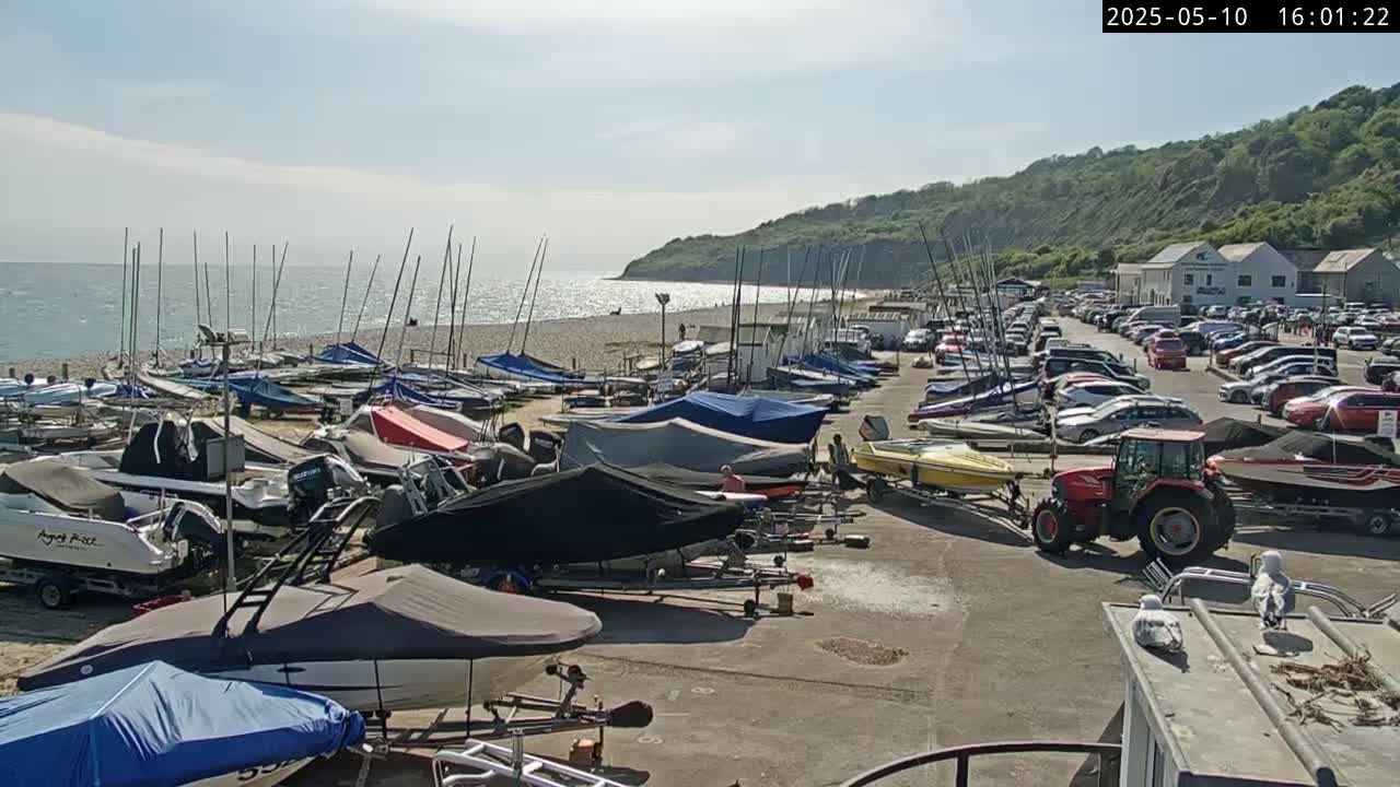 Many boats and sailboats are parked on a sandy beach next to a parking lot full of cars under a sunny sky.