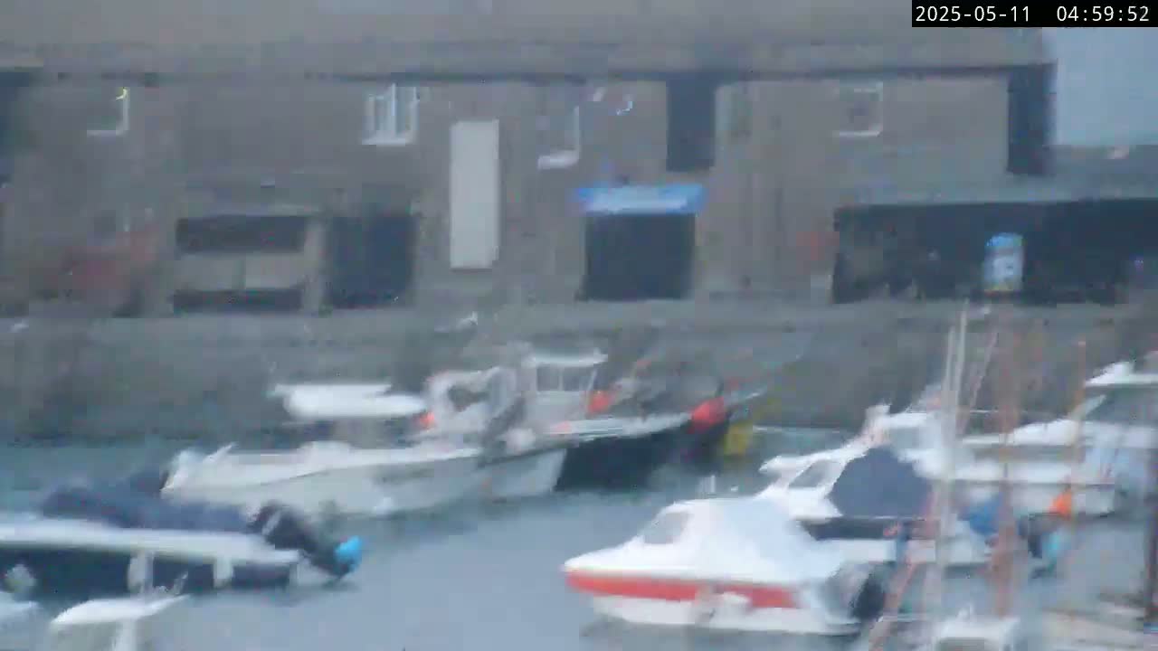 Several small motorboats are moored in a harbor next to a stone building on an overcast day.