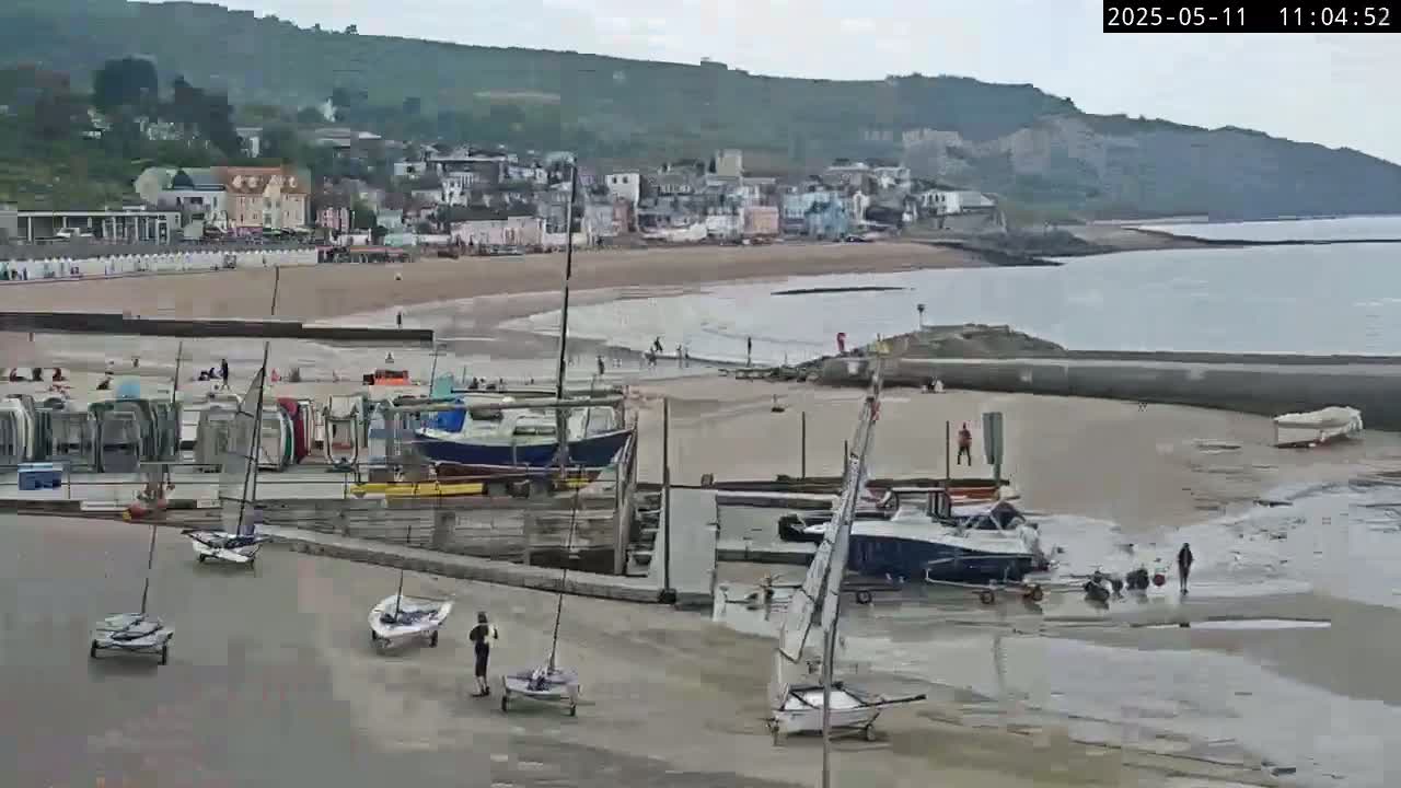 A seaside town with a sandy beach, several small sailboats on land, and a few people scattered about, under overcast skies.