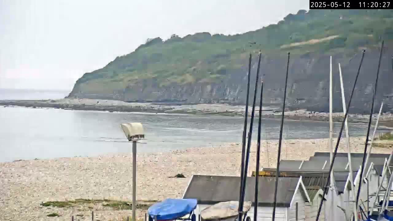 A pebble beach with several small beach huts and sailboat masts is visible in the foreground, with calm water and a green cliff in the background under an overcast sky.