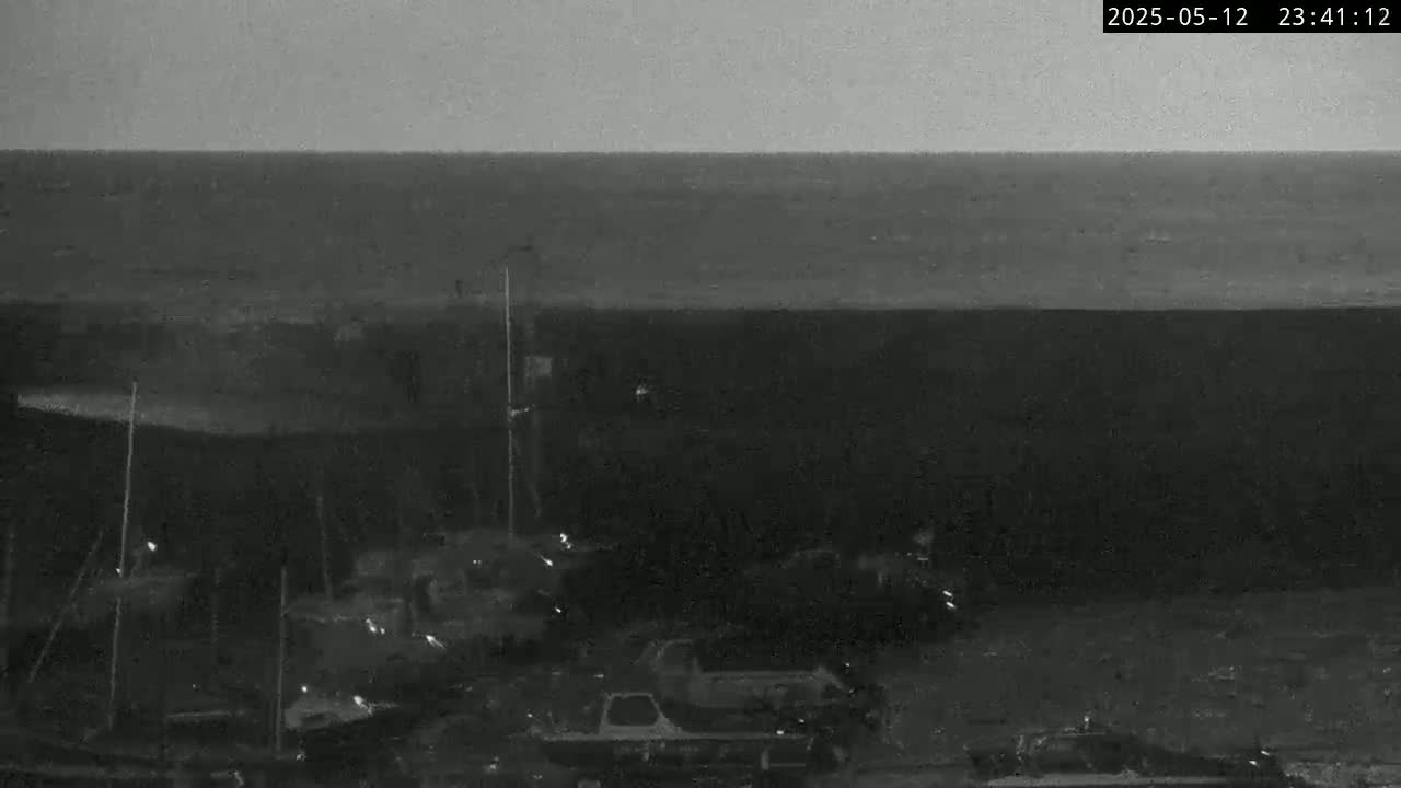 A nighttime, low-light image shows a dark body of water, possibly the ocean, with a harbor or marina containing several boats in the foreground under an overcast sky.