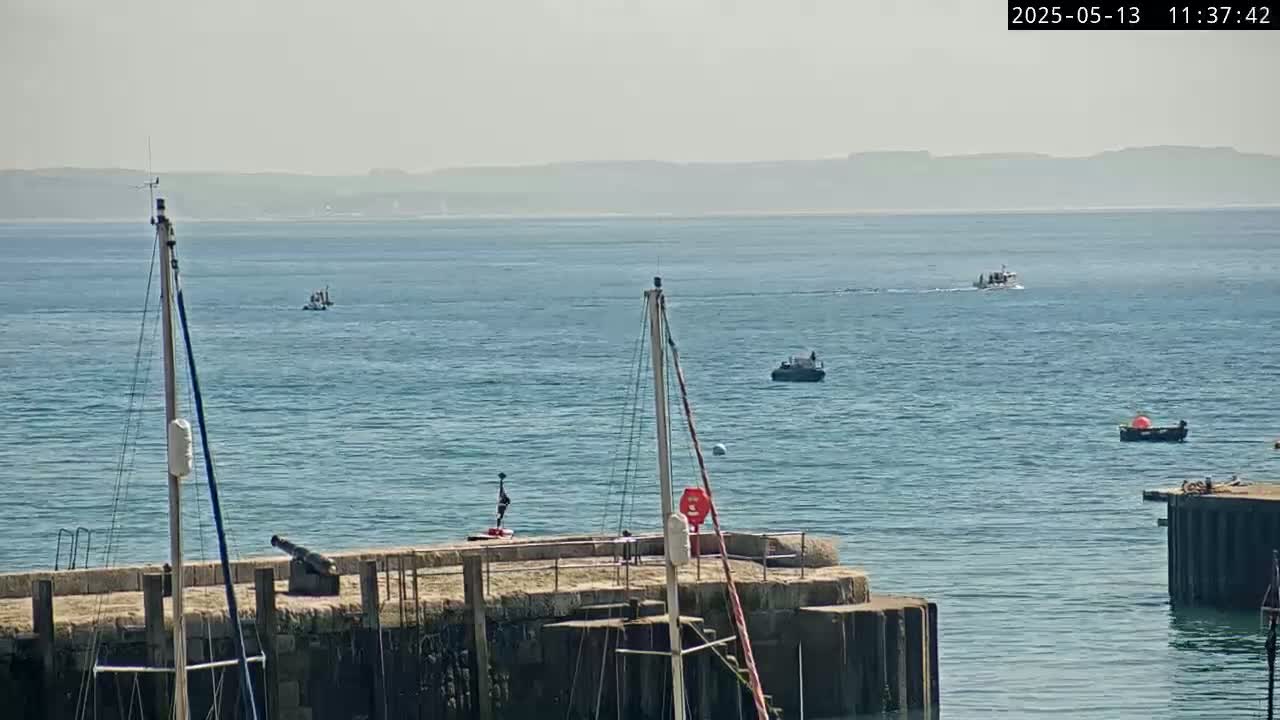 Several boats are on a calm, hazy sea near a stone pier with masts and a cannon.