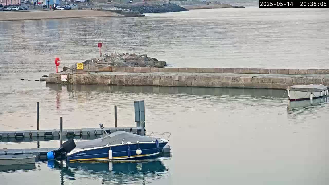 A calm body of water shows a small motorboat docked at a stone jetty, with another boat further out, under a cloudy sky.