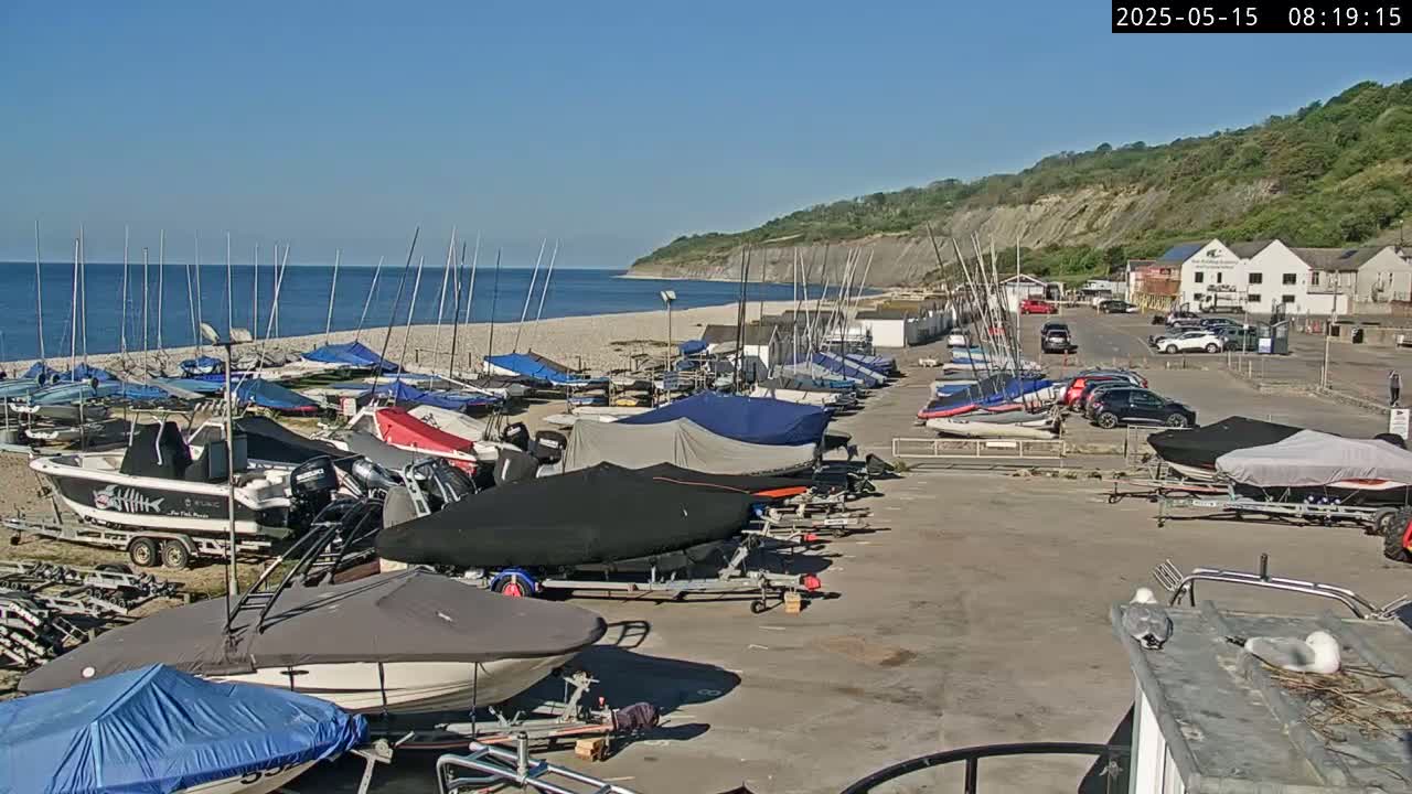 Numerous boats and sailboats are parked in a lot next to a beach and a cliffside on a sunny day.