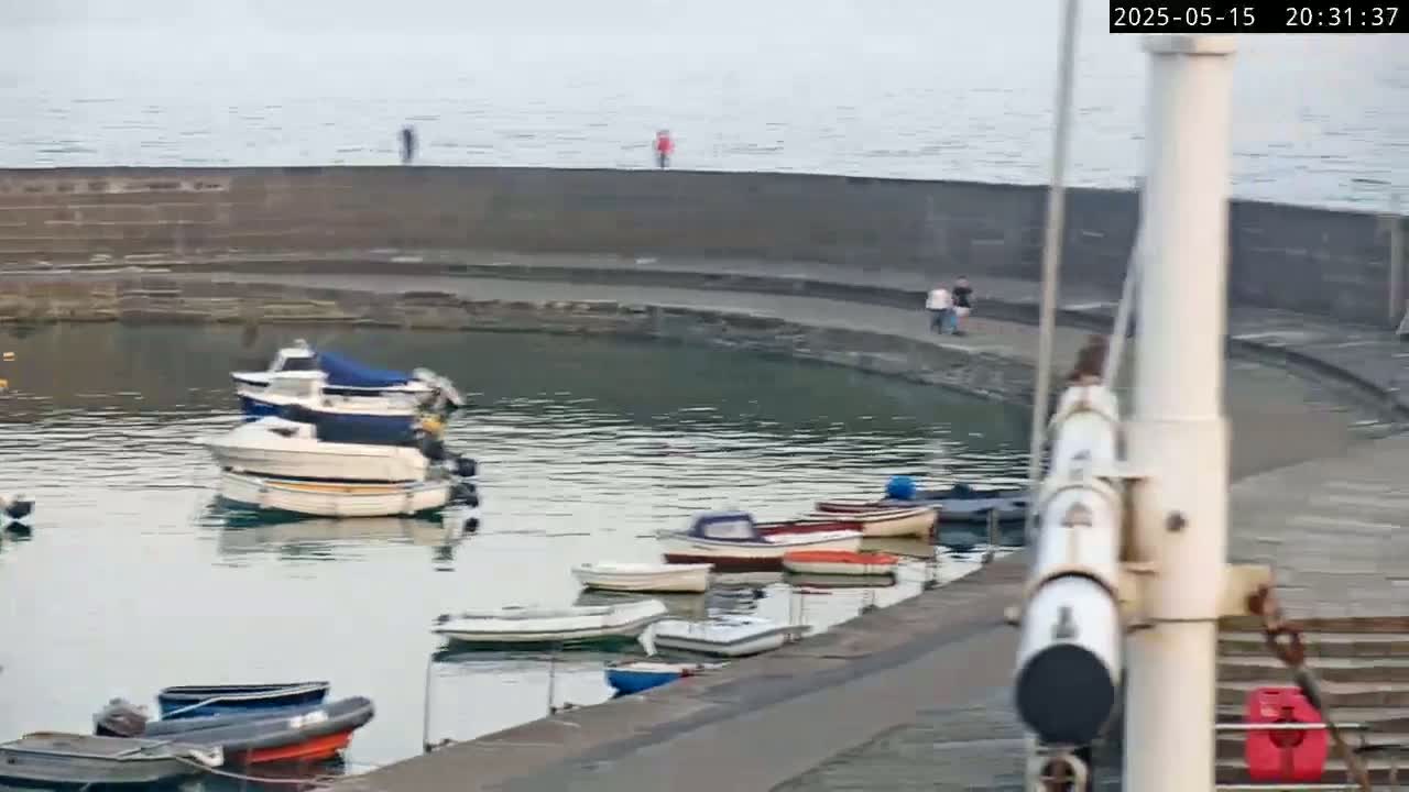 Several small boats are moored in a calm harbor, next to a stone wall where a few people are walking, under a cloudy sky.