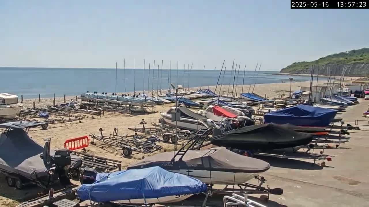 Numerous boats and sailboats are parked on a sandy beach next to a calm ocean under a sunny sky.