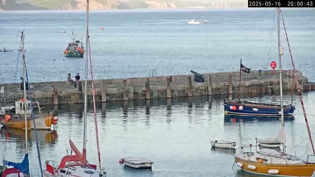 Several sailboats and small fishing boats are moored in a calm harbor next to a stone pier with a cannon, under a clear evening sky.
