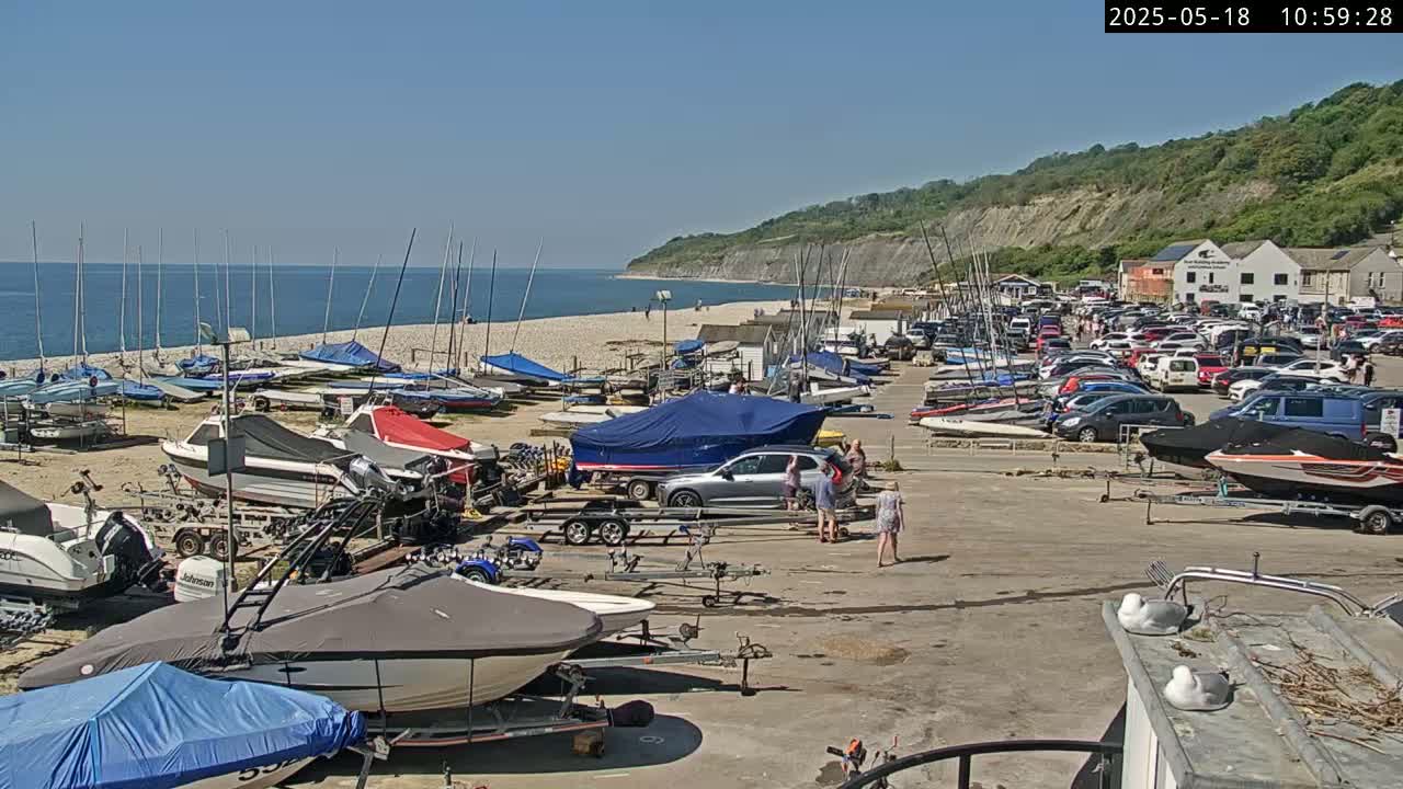 A sunny day at a beach with numerous sailboats and motorboats docked on a pebble beach near a parking lot full of cars and a cliff in the background.