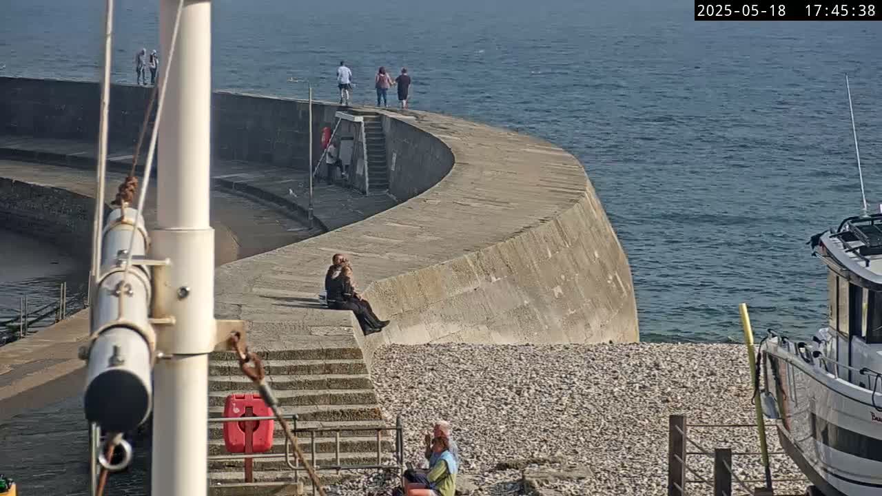 A curved concrete seawall with several people sitting and standing on it overlooks a pebble beach and calm ocean under a sunny sky.