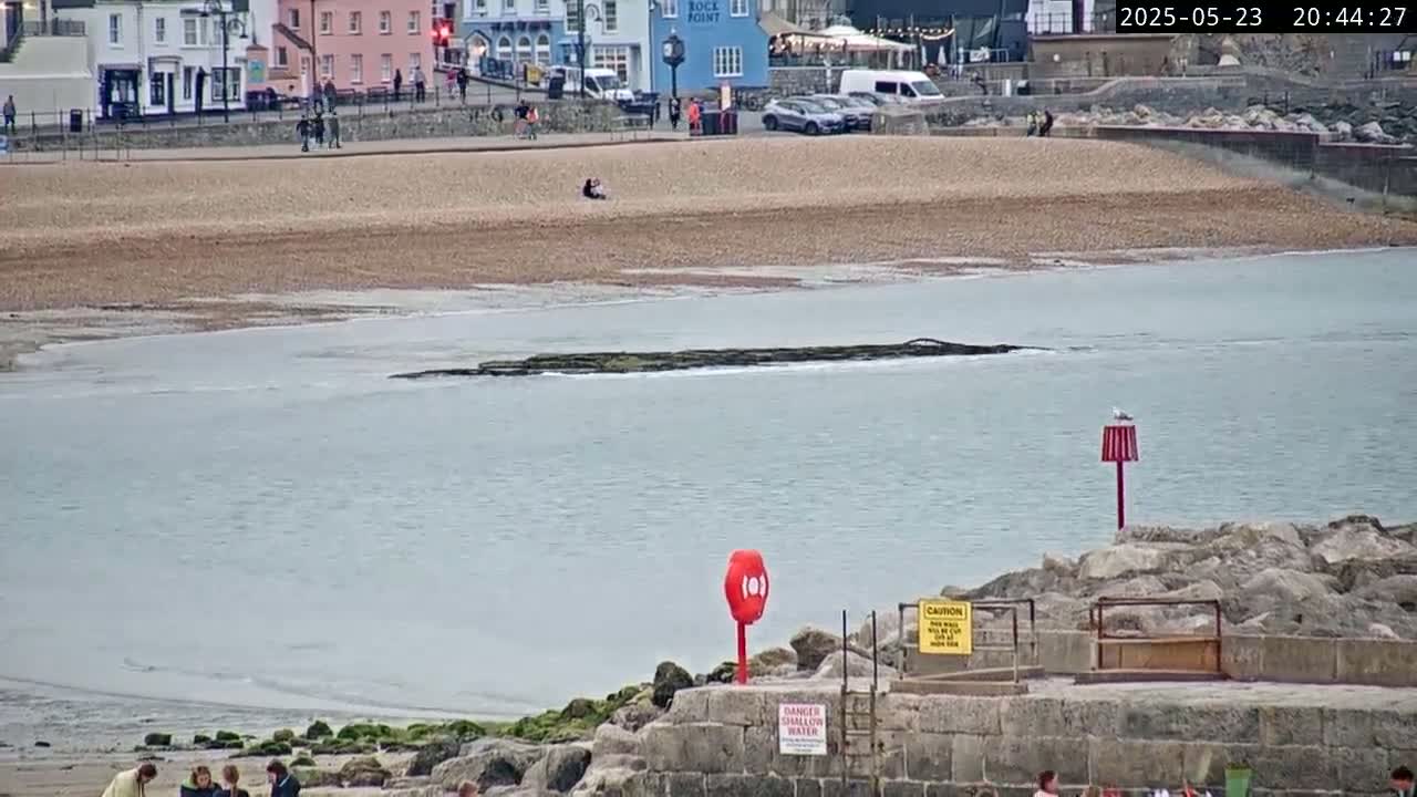 A calm sea laps a pebble beach near a stone seawall, with buildings and a few people visible in the background.