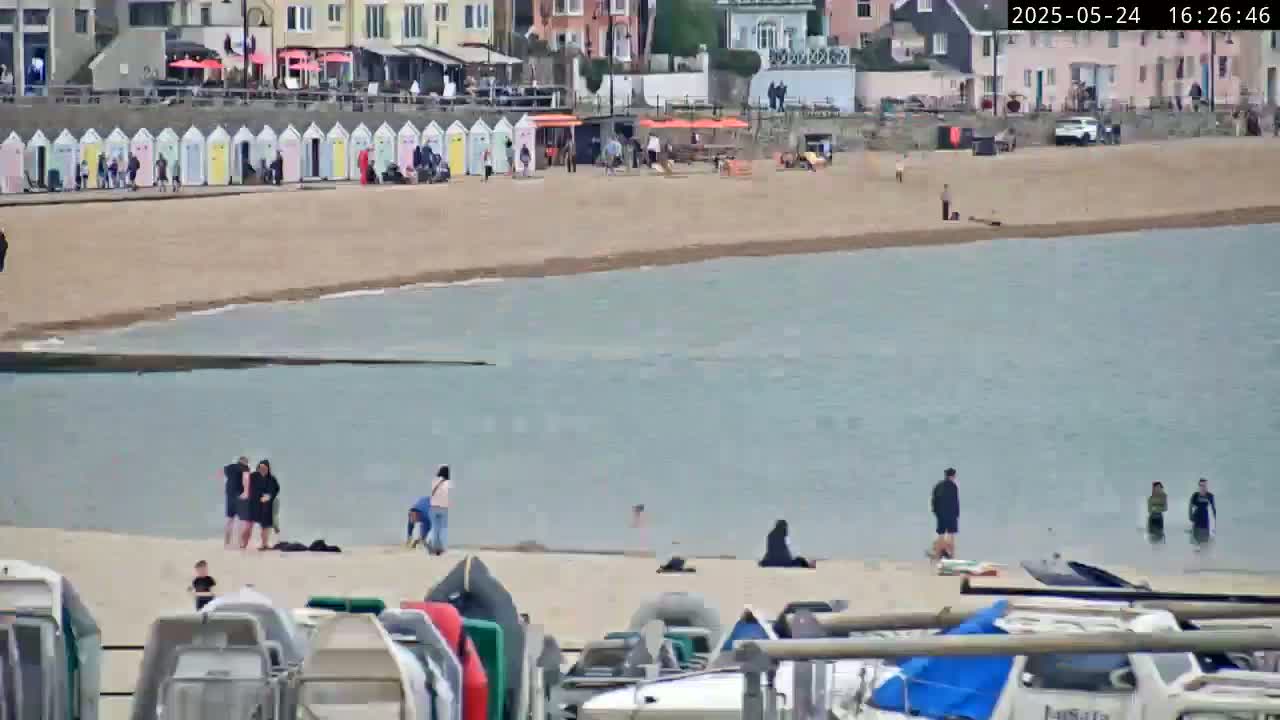 A sandy beach with calm, light-blue water, several people scattered on the sand and in the water, and numerous boats pulled up on shore.