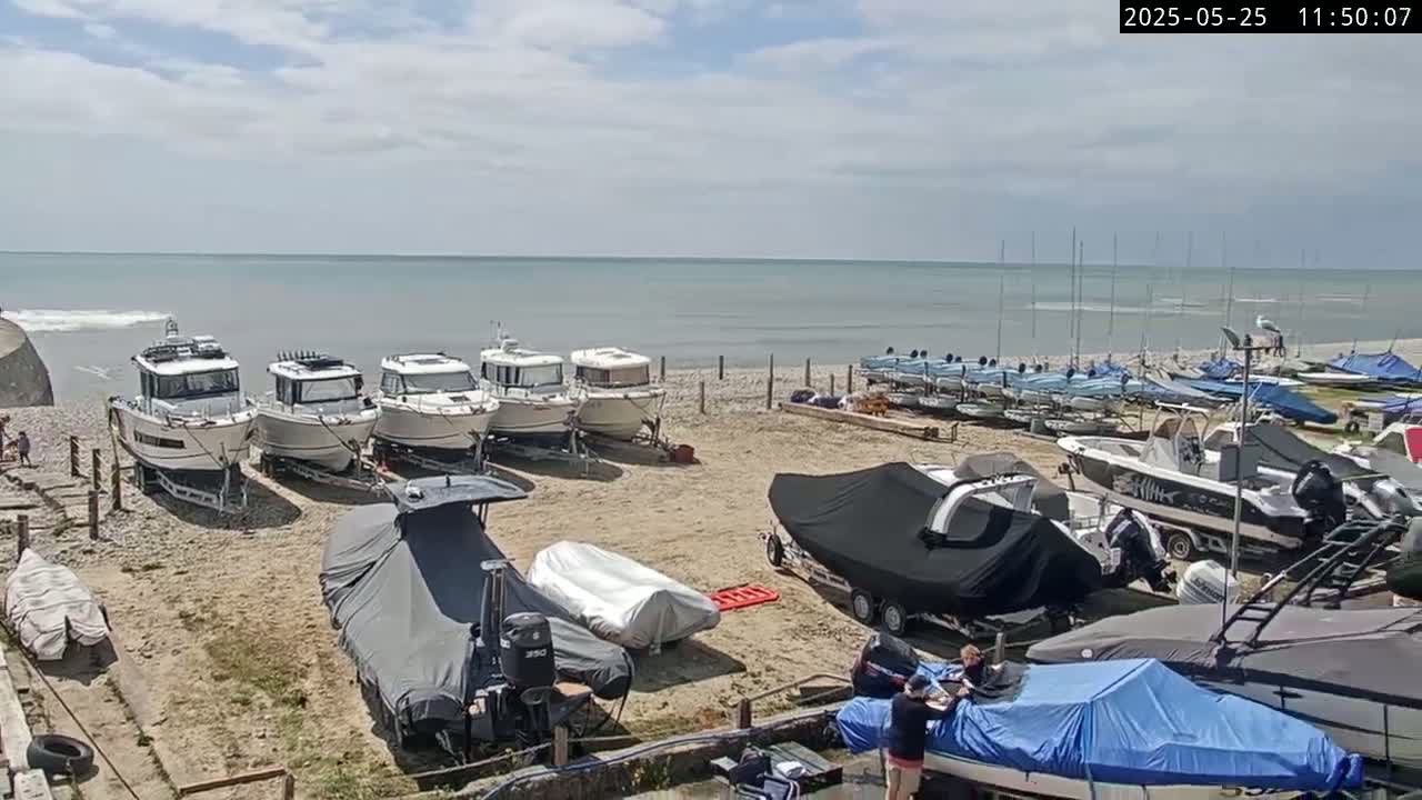 Several boats are parked on a pebble beach next to a calm ocean under a partly cloudy sky.