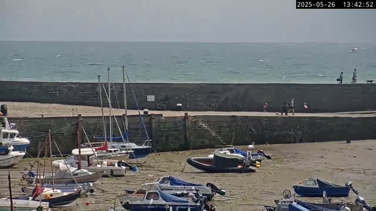 A harbor filled with small boats sits on a sandy shore next to a stone wall, with people walking along the wall overlooking a calm, windy ocean.