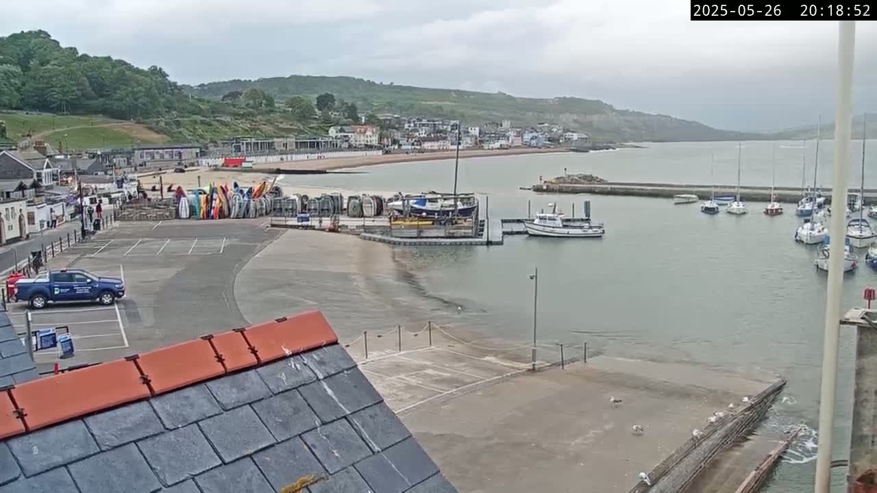 A harbor scene on an overcast day shows a small town nestled against a hillside overlooking a calm bay with several boats moored in the harbor and a few parked vehicles visible.