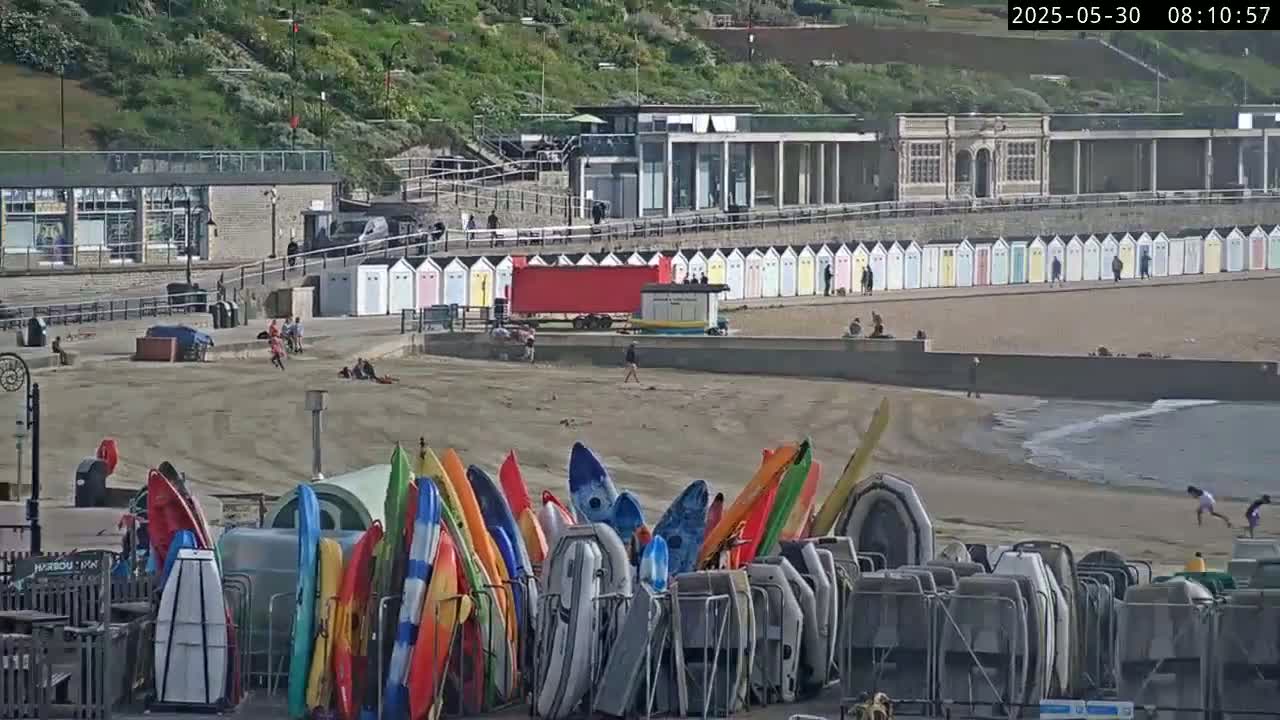 A sandy beach with colorful kayaks and beach huts is seen on a partly sunny day, with a calm ocean in the background.