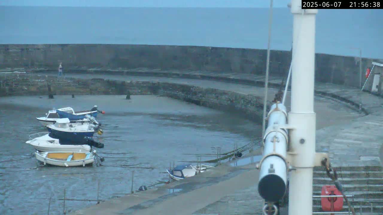 Several small motorboats are moored in a calm harbor at low tide, next to a stone wall under an overcast sky.