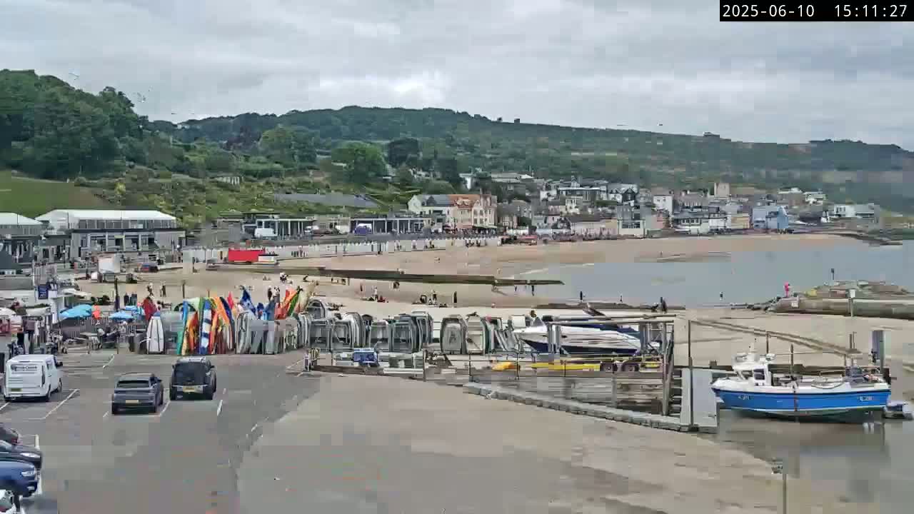A beach scene on an overcast day shows a sandy beach with people, beach huts, boats, and a parking lot with cars.