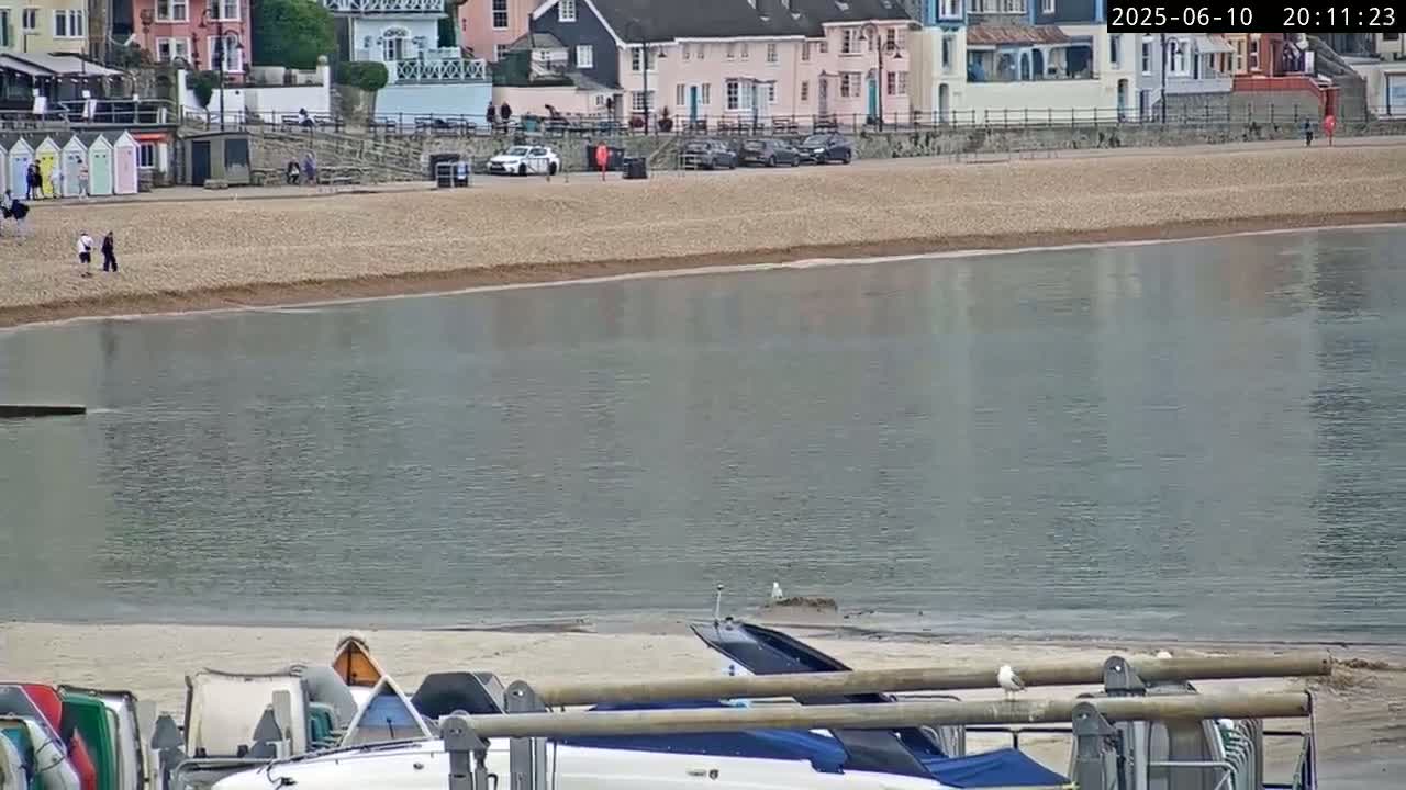 A calm body of water laps a pebble beach in front of colorful buildings; several parked cars and a few people are visible on the beach.
