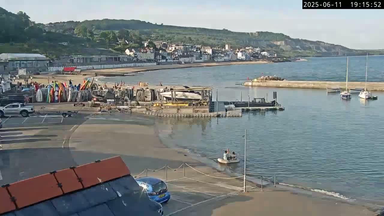 A seaside town with a sandy beach, a small harbor containing several boats, and a calm sea under a clear sky.