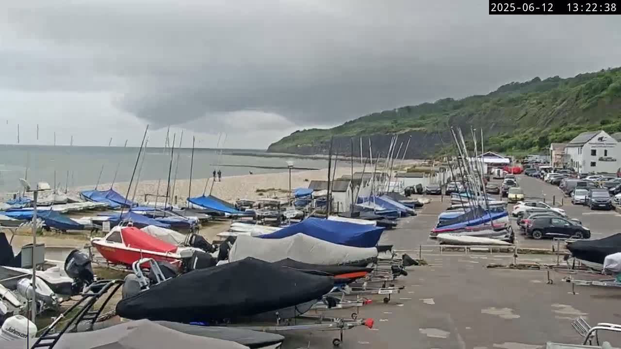 Numerous sailboats and motorboats are docked at a harbor next to a sandy beach, backed by a steep, grassy cliff under an overcast sky.