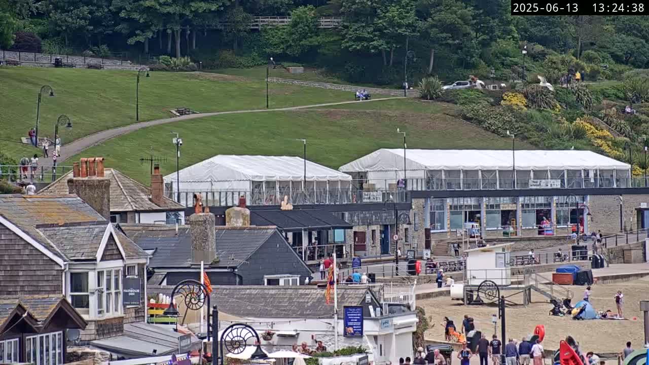 A seaside town on a sunny day, with people on a beach below buildings and grassy hill with a walkway above.