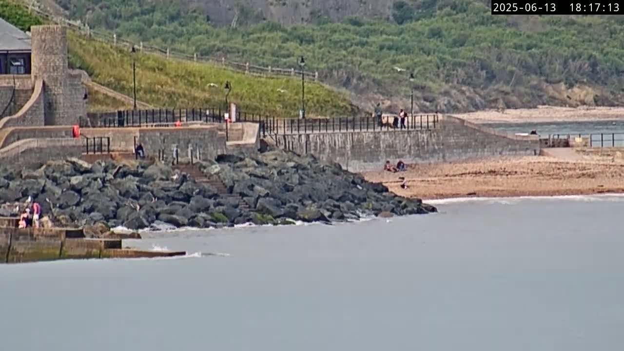 A rocky shoreline with a few people on a sandy beach, a stone wall with a walkway above, and a building in the background on a partly cloudy day.
