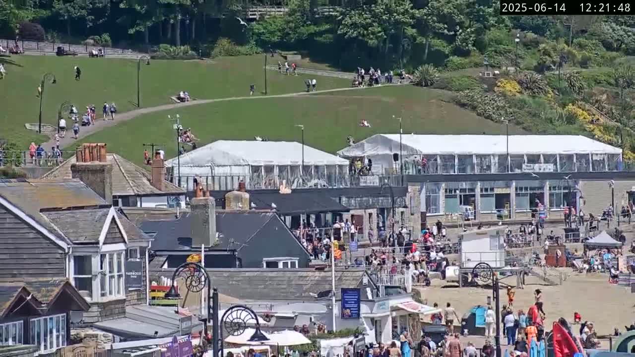 A sunny seaside town scene shows a grassy hill with people walking, two large white tents near a building, and a crowded beach with people relaxing and enjoying the day.