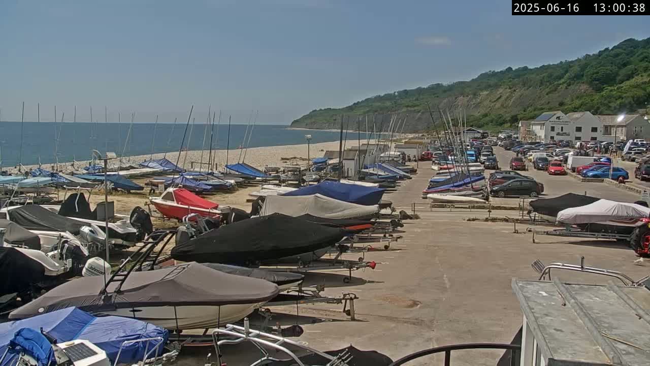 Numerous boats are parked in a lot next to a sandy beach, with a calm ocean and green hills in the background under sunny skies.