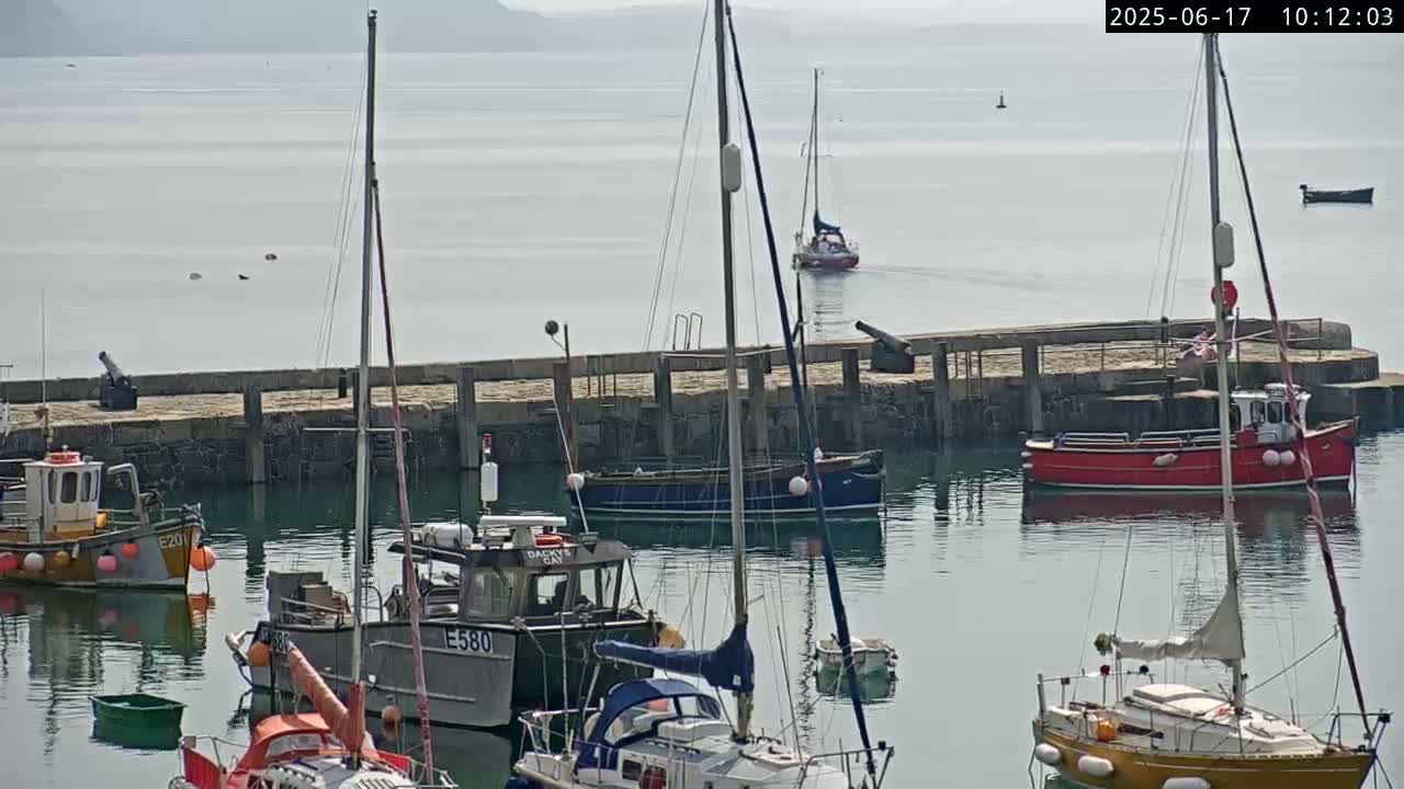Several boats are moored in a calm harbor on a hazy day, with a pier separating them from the open water.