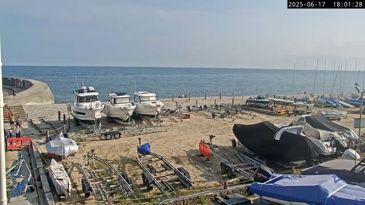 Several boats on trailers are parked on a pebble beach next to a calm sea on a sunny day.