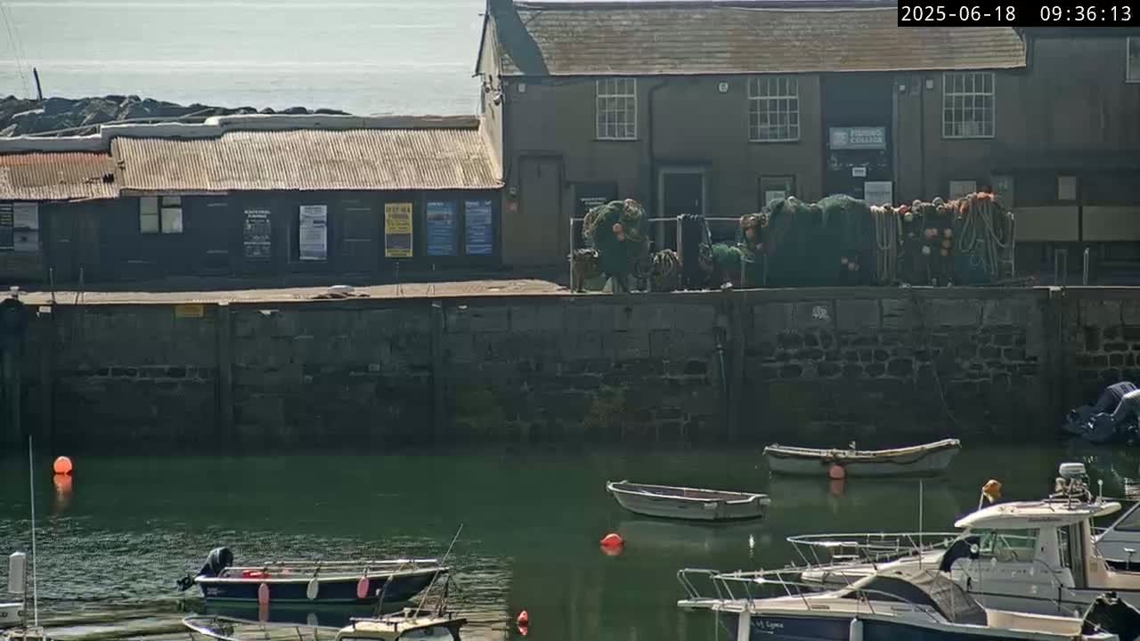 A calm harbor scene on a sunny day shows several small boats moored near a stone wall and buildings, with fishing nets piled on top of the wall.