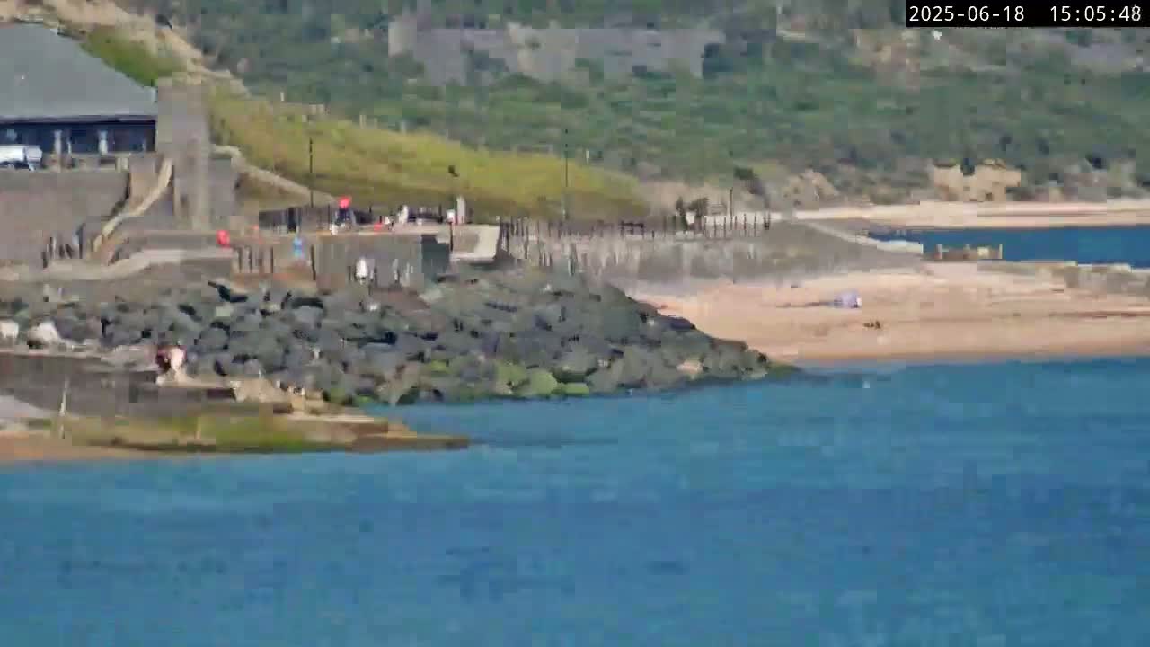 A sandy beach with a few people on it is next to a rocky shoreline, with a building and paved walkway visible in the background on a sunny day.