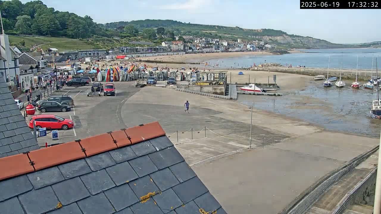 A sunny day overlooking a coastal town's harbor, with a sandy beach, parked cars, boats in the harbor, and a person running in a paved area.