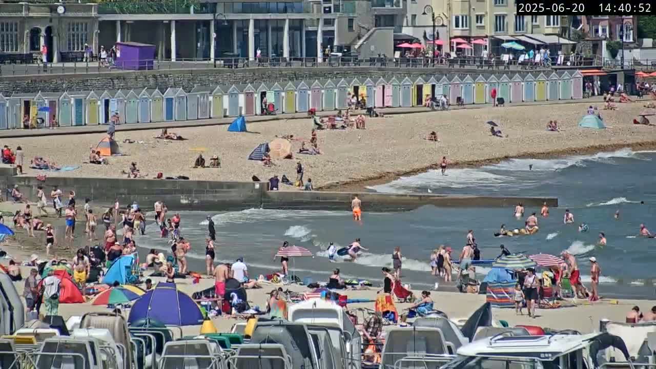 A sunny beach scene shows many people relaxing, swimming, and sunbathing on a pebbled beach, with small boats and beach huts in the background.