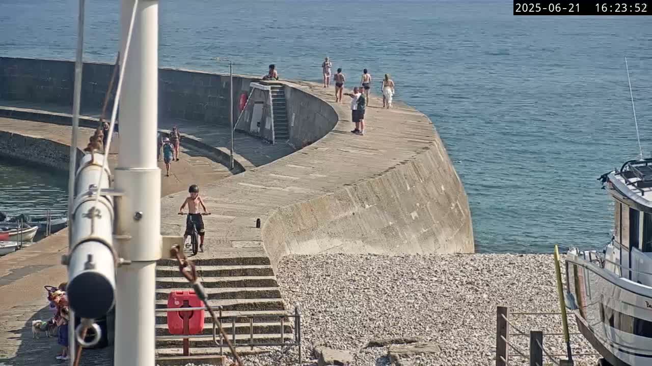 A curved concrete wall extends into a calm sea on a sunny day, with several people walking along it and a child riding a bicycle near a pebble beach.